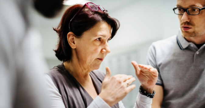 A female manager with shoulder-length dark hair uses hand gestures in a conversation with her team, displaying purpose-driven leadership through her communication style