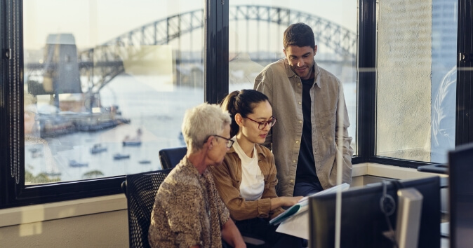 A group of three employees engaged in a discussion about team dynamics in an office setting. Two women, one with gray hair and the other with long hair in a ponytail, are seated while a man stands beside them, looking at a document held by the woman with the ponytail.