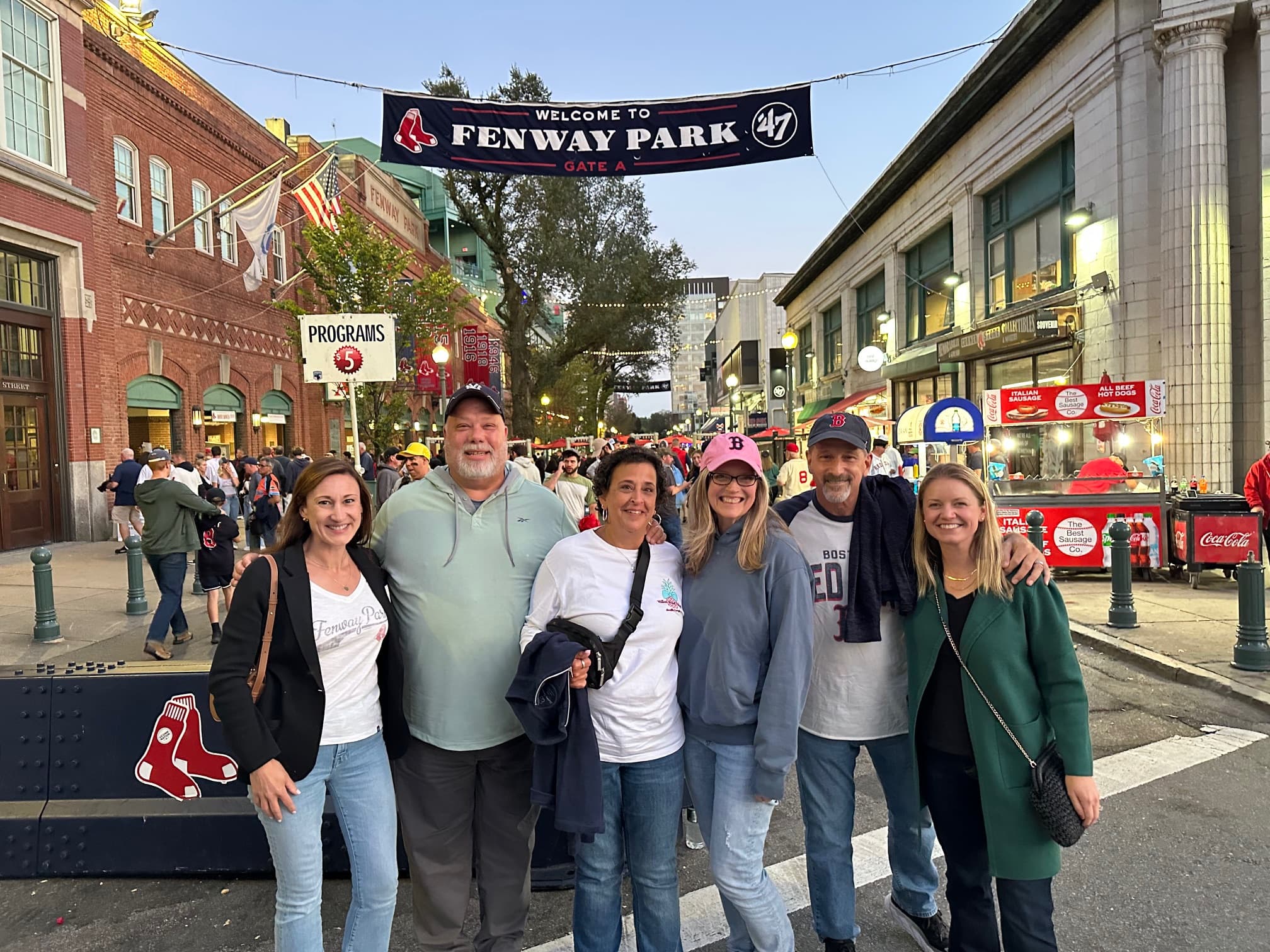 A group of six people smiles in front of a "Welcome to Fenway Park" banner. The street is bustling with people and baseball atmosphere.