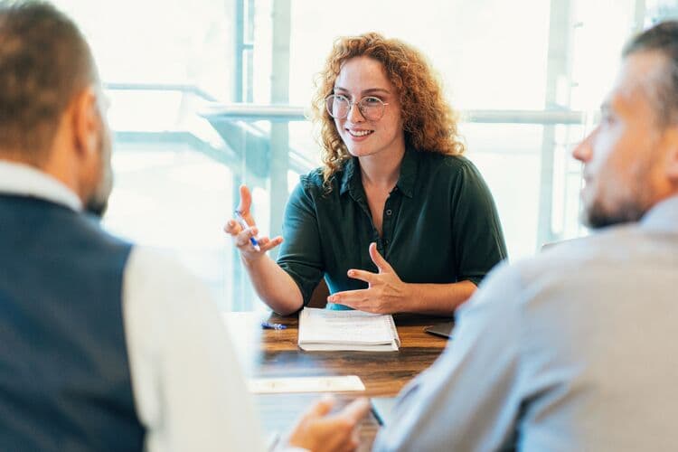 A woman, who is a part of an adhocracy culture, is engaged in a conversation at a meeting table, using expressive hand gestures. She is smiling and appears to be discussing ideas with two men whose faces are partially visible. There are notes and a smartphone on the table, and large windows in the background let in natural light.